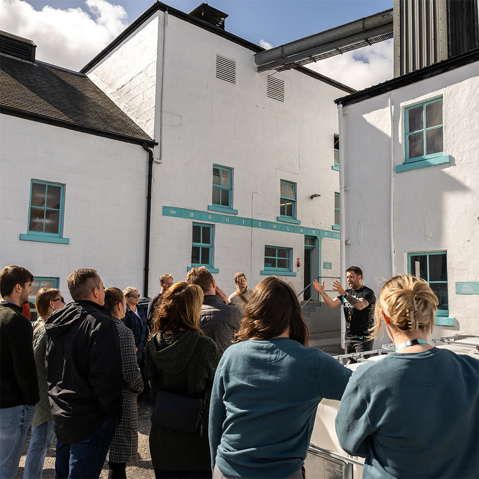 A group of visitors listening to a tour guide at the Bruichladdich Distillery courtyard, with the iconic whitewashed buildings and aqua accents under a sunny sky.