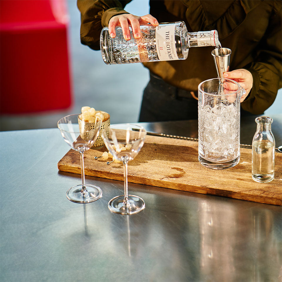 A person preparing a cocktail with The Botanist gin, pouring it into a mixing glass on a wooden board, accompanied by garnished glasses and ingredients.