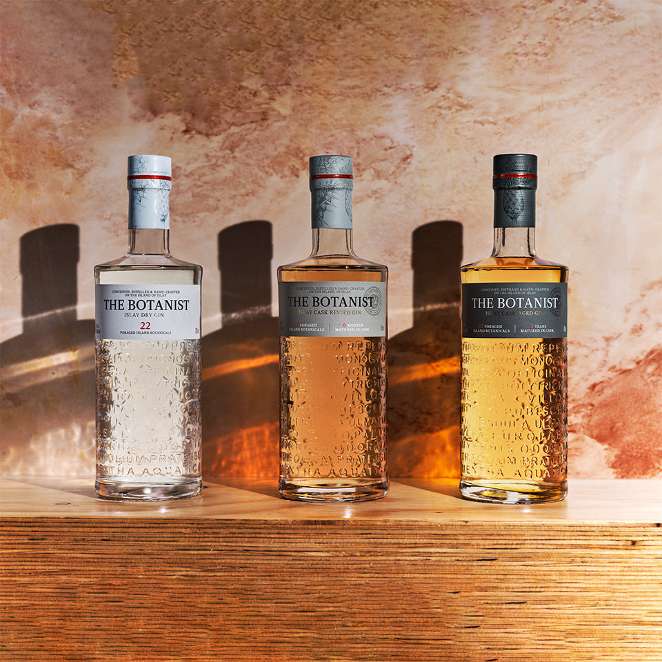 Three bottles of The Botanist gin displayed on a wooden surface against a warm, textured background, showcasing their clear and cask-aged expressions.