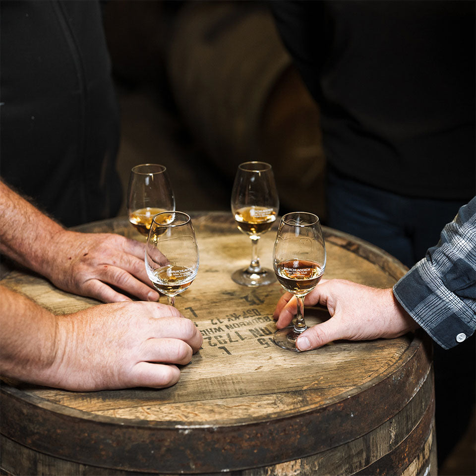 Three hands resting on a whisky barrel, each holding a glass of whisky during a Bruichladdich Distillery warehouse tasting.